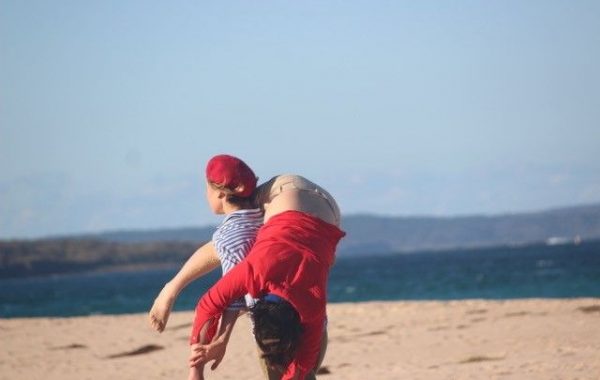 A woman in a red beret looks out to sea on a beach. She is carrying a person in a red jumper over one shoulder