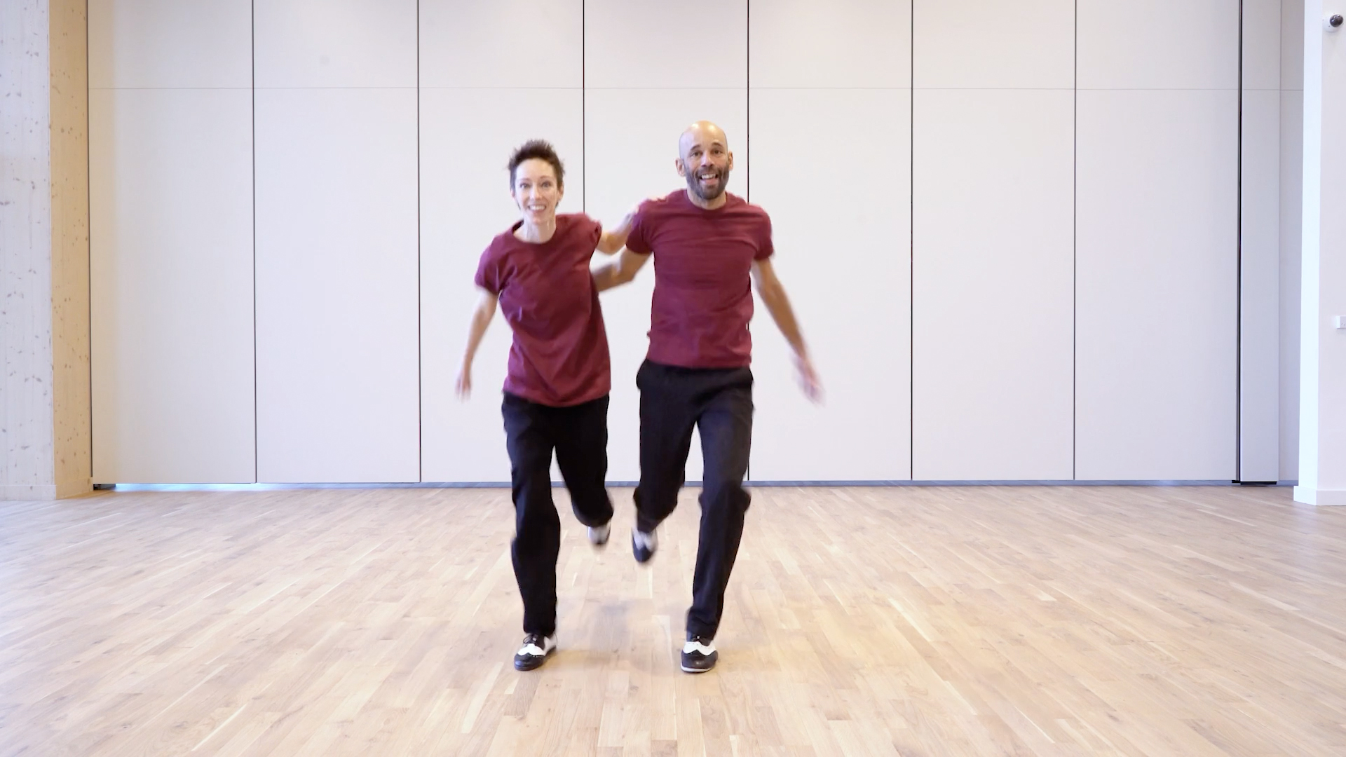 Lead & Follow Partner Work. Dance teachers Wendy Steatham, a short white woman with brown spiky hair, and Temujin Gill, a tall man with light brown skin and a shaved head, are dancing with one foot off the floor. Wendy's hand is on Tem's shoulder