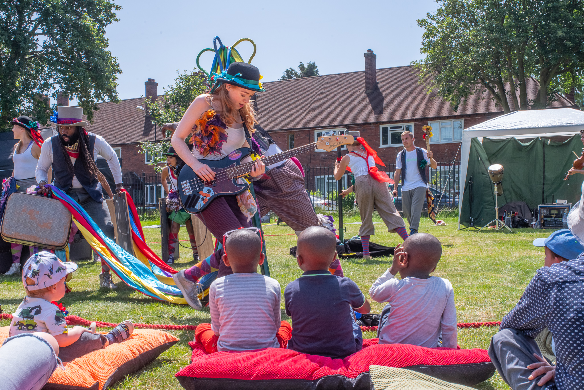 Folk Dance Remixed. A group is performing to some children in a park. The most prominent figure holds an electric guitar and is wearing a top hat. There are three children with their backs to us, sitting on a large red cushion