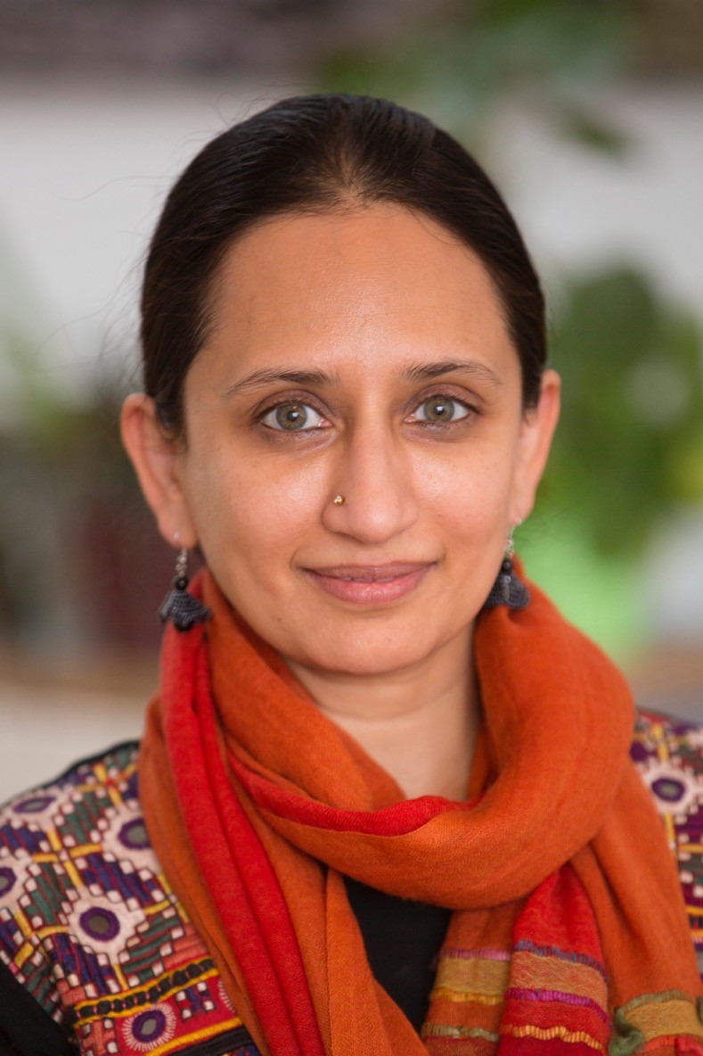 Anusha Subramanyam. A head shot of a smiling woman with dark, centre parted hair and light brown eyes. She has a nose stud and is wearing drop earrings and a bright orange scarf