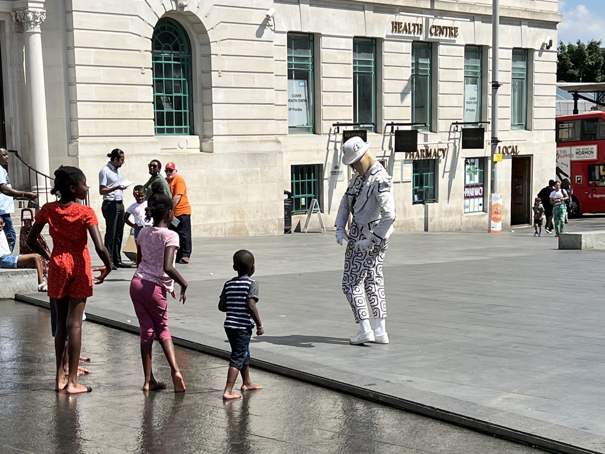 The RIDDLE in General Gordon Square. A costumed character poses in front of a group of children. He wears a white suit with a white bowler hat