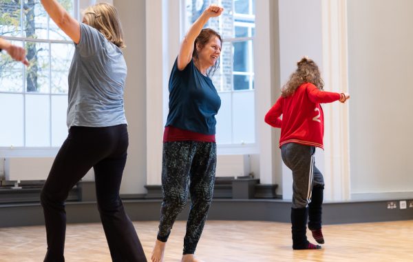Dance for Wellbeing. Three dancers are in a light studio. The central dancer is raising an arm like she is punching the air. She is smiling. We cannot see the faces of the other two as they are turned away from the camera