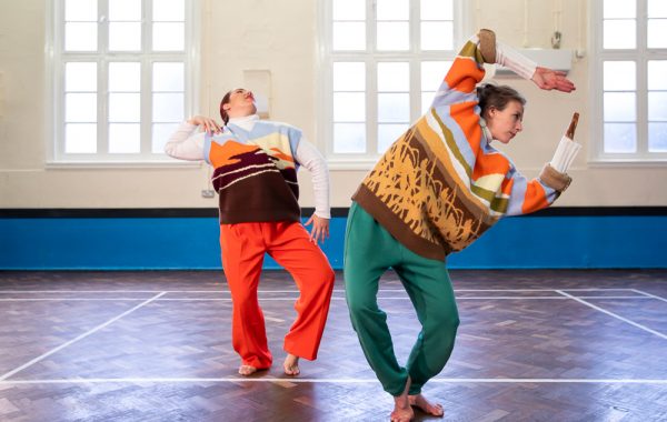 Two performers in bright coloured trousers and knitted jumpers pose in a studio with wooden floors