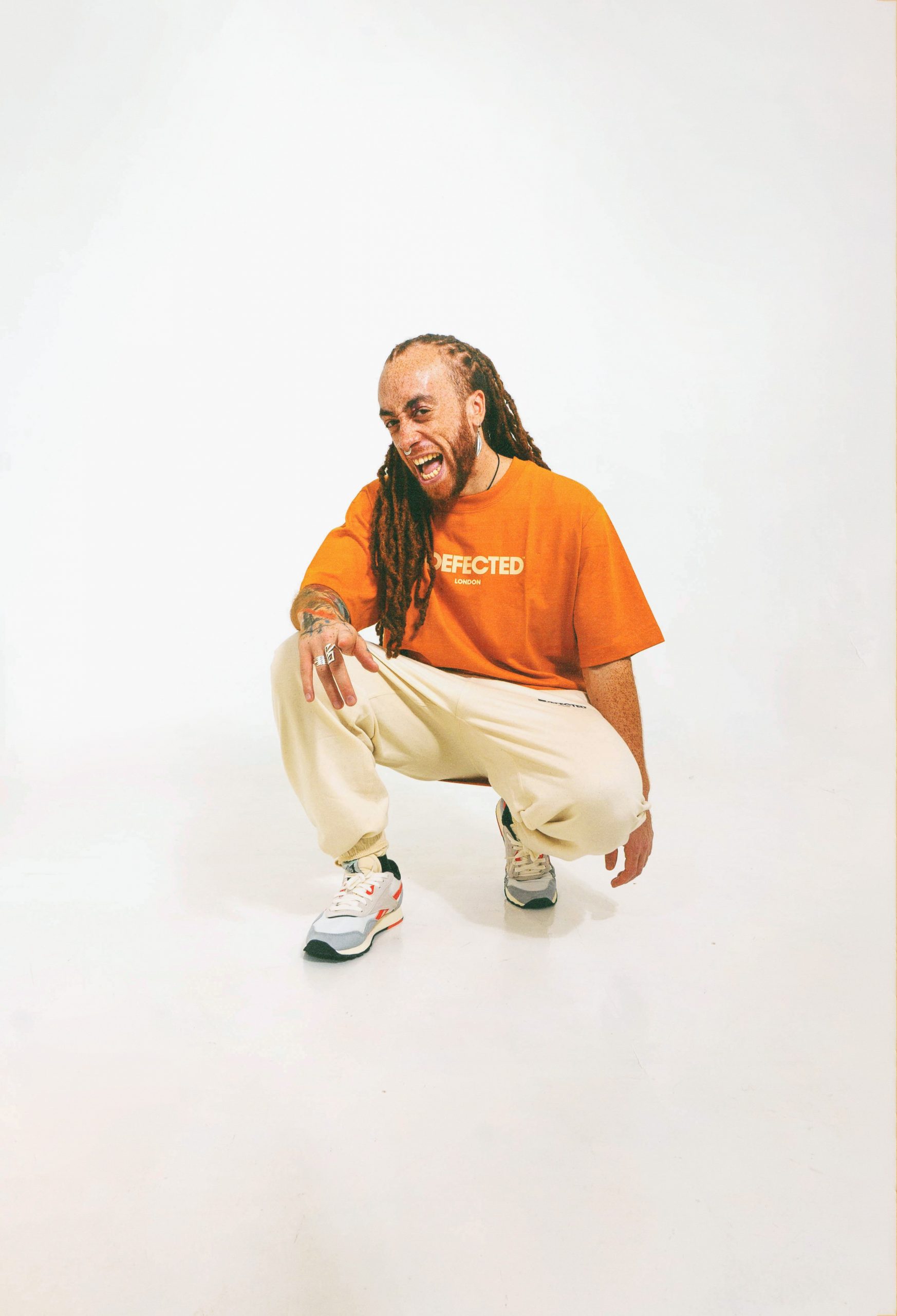 image of Frankie. Frankie is wearing a bright orange top with white trouser and his distinctive long red dreads. He is crouching down in front of a white background. Credit to @minaenea