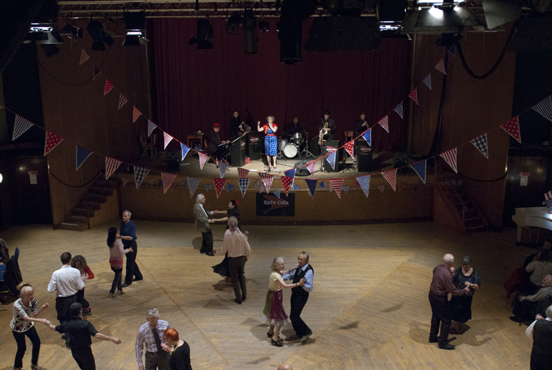 A tea dance in the Borough Hall. There is a band on stage and bunting is arranged around them. Couples dance on the wooden floor.