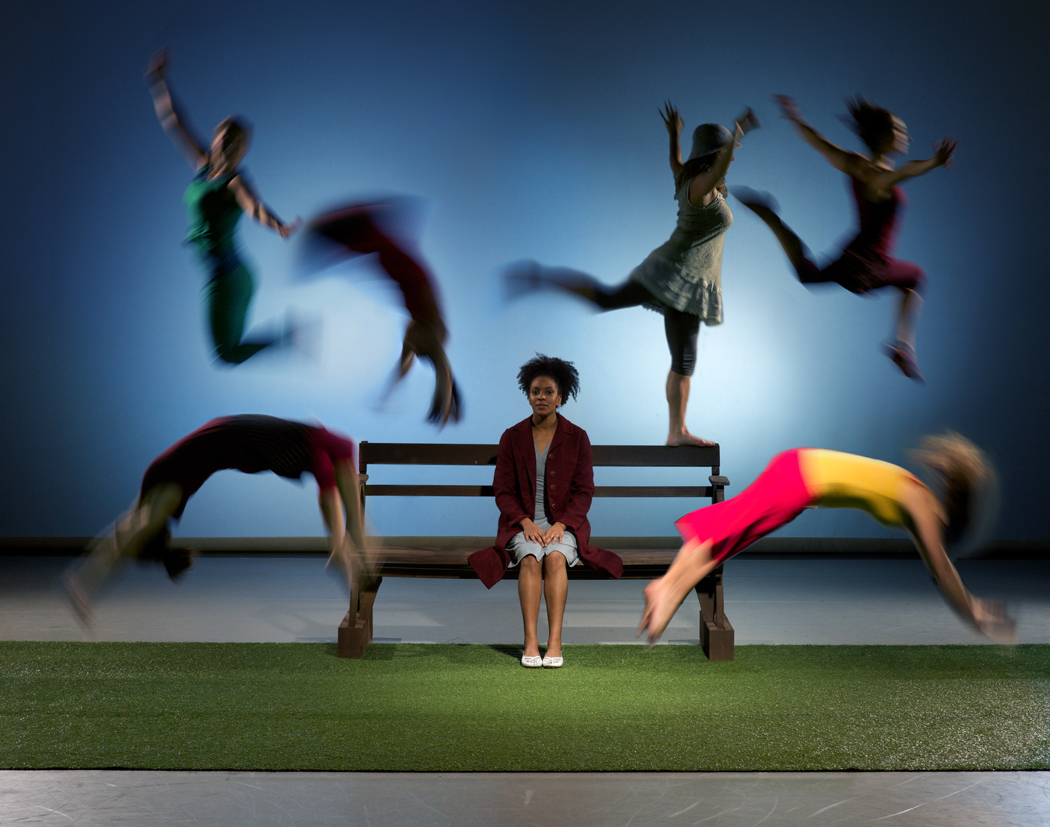 30 years in pictures: Compass Commissions Part 1. A woman is sat on a park bench and six performers leap from the bench all around her