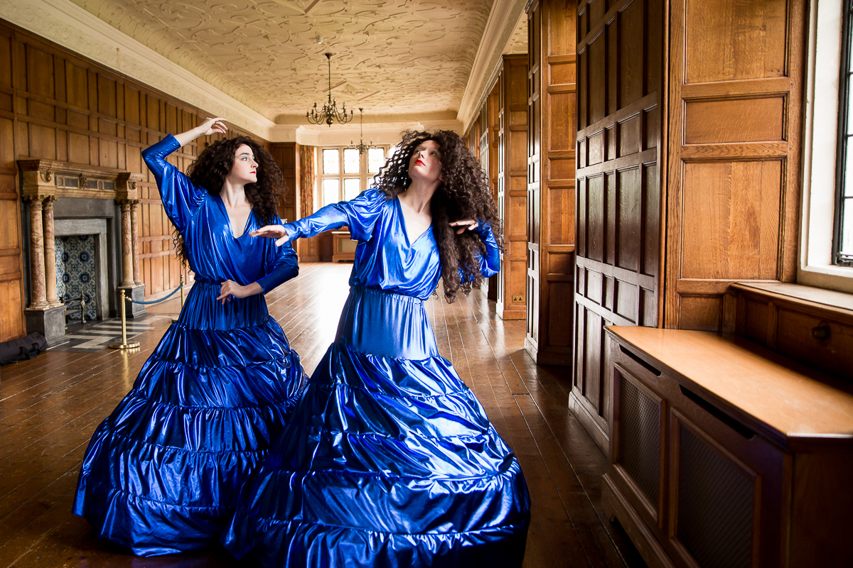 30 years in pictures: GLORIA! and Charlton House. Two dancers in bright blue hooped dresses pose in the long gallery, a wood panelled room at Charlton House.