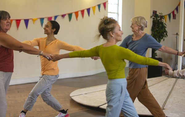 Image shows four adults dancing and holding hands as they perform in a community hall to an audience. There is bunting behind and the dancers smile as they move. Image credit, Kinga Dawid