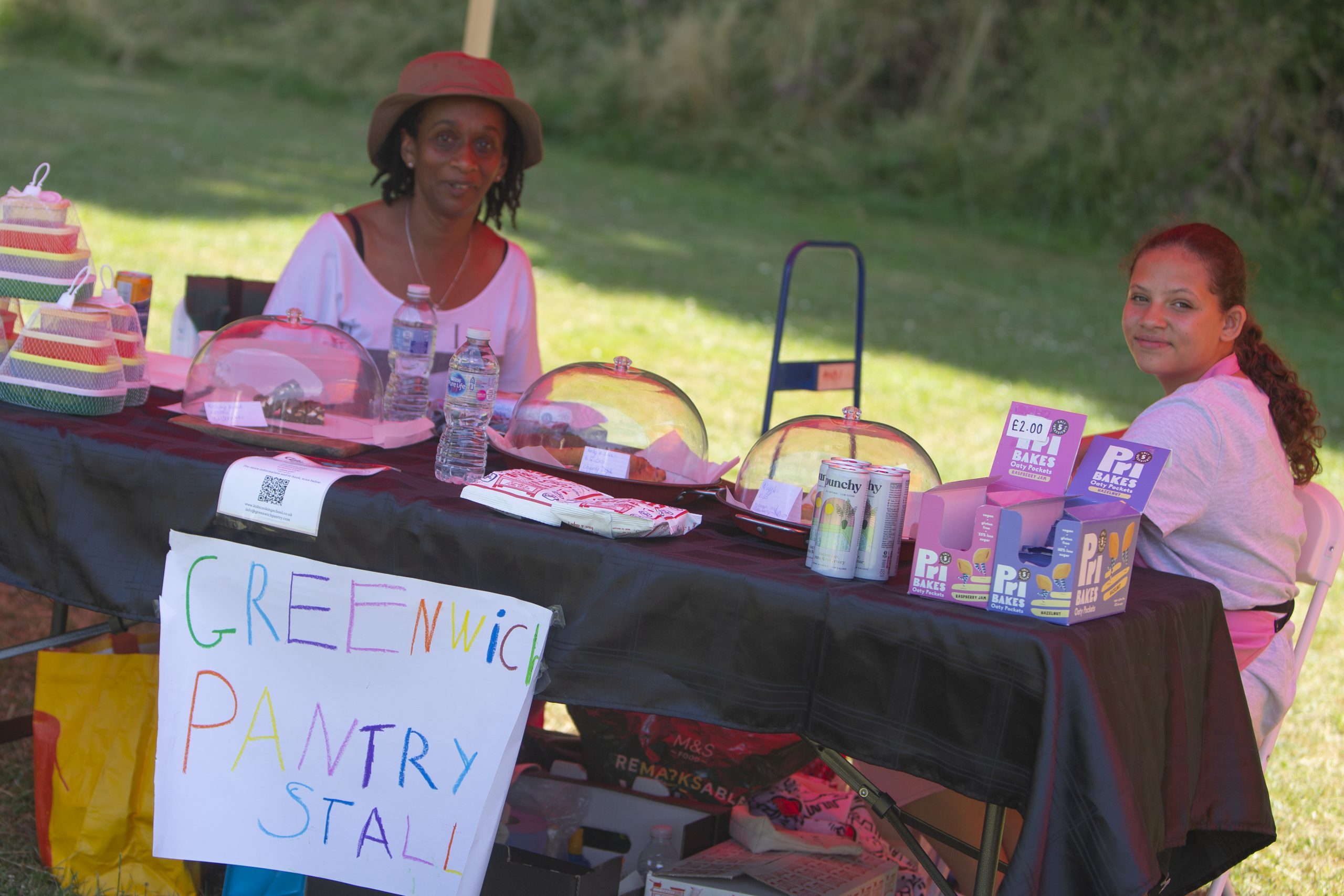 Remembering a glorious Summer in the Park. Two people from the Greenwich Pantry sit behind their display of cakes and their hand written sign which reads Greenwich Pantry Stall.