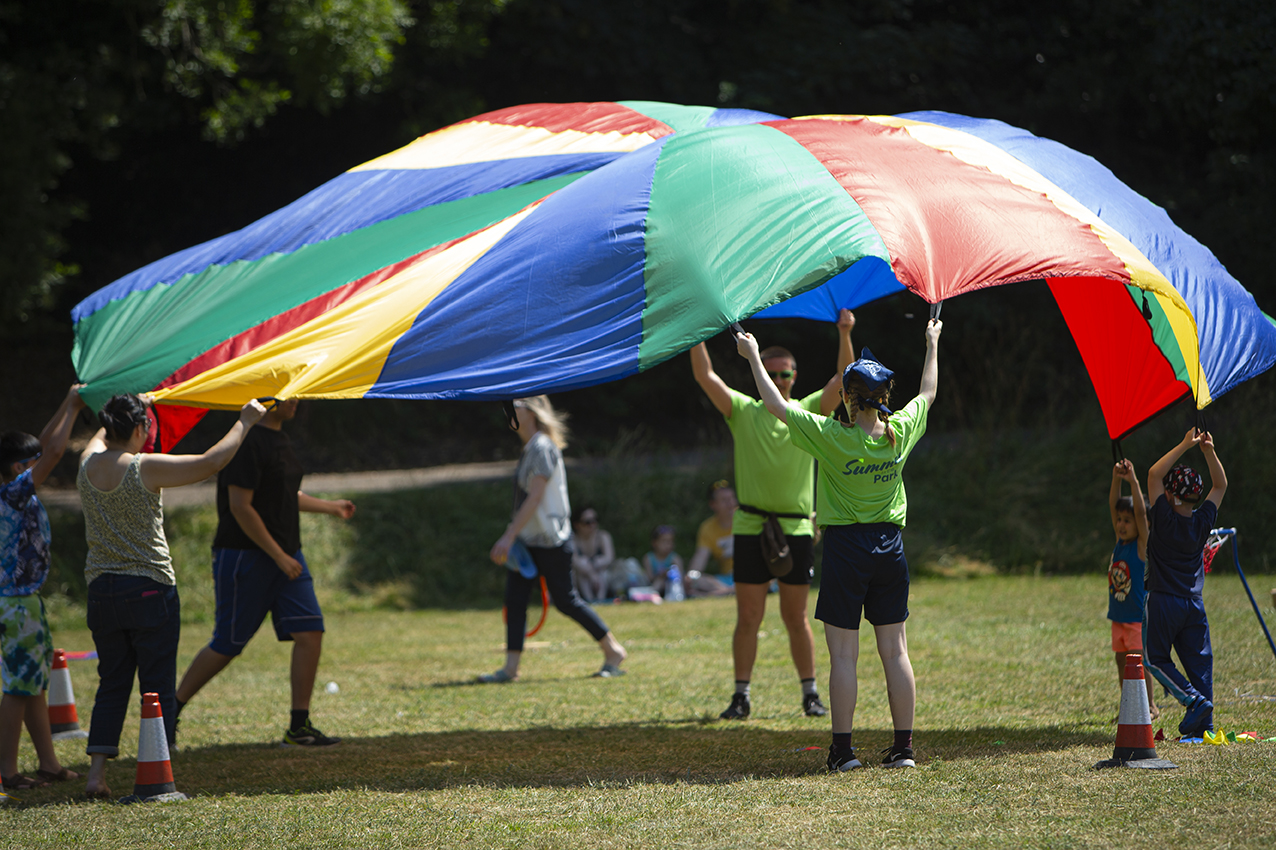 Remembering a glorious Summer in the Park. Several people are in a park and holding a large colourful parachute aloft.