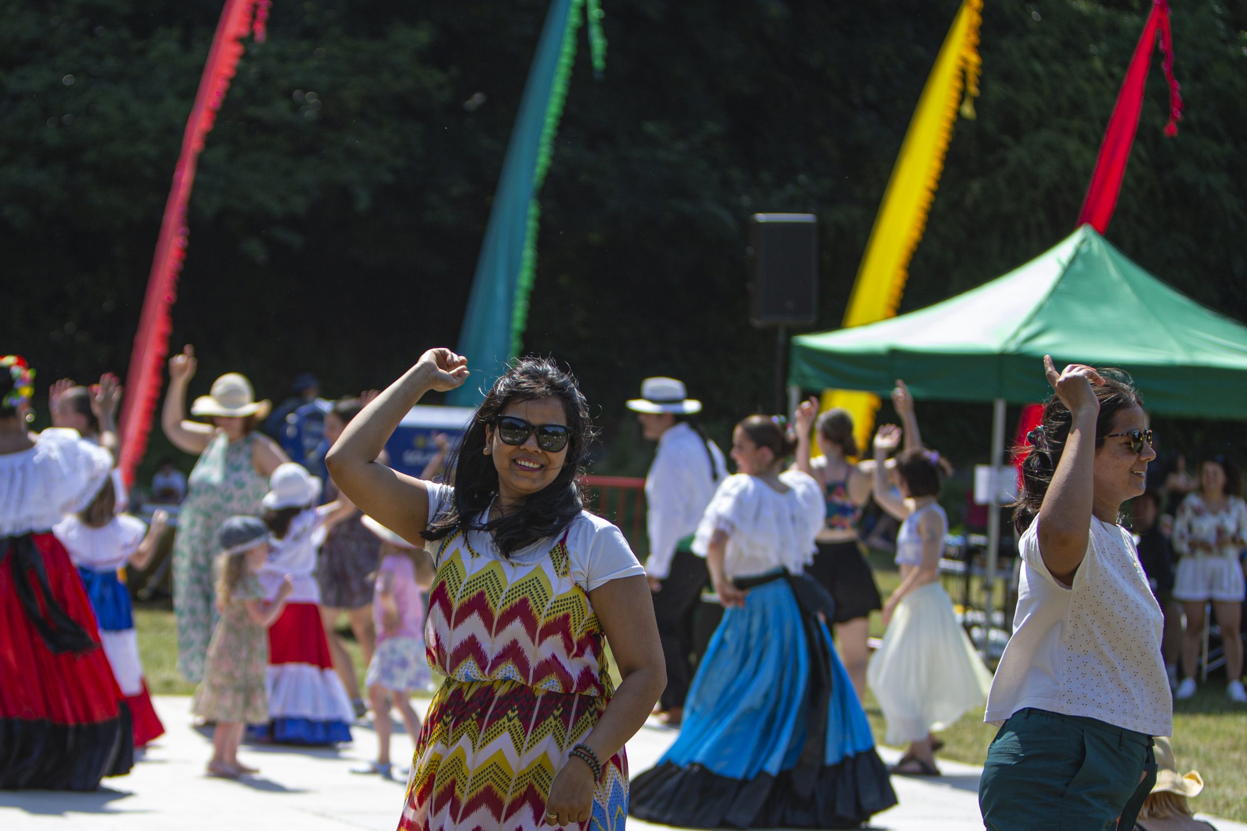 Remembering a glorious Summer in the Park. An outdoor event is taking place with dancers in colourful skirts twirling on a flat stage. In front, an audience member is smiling and joining in the fun.