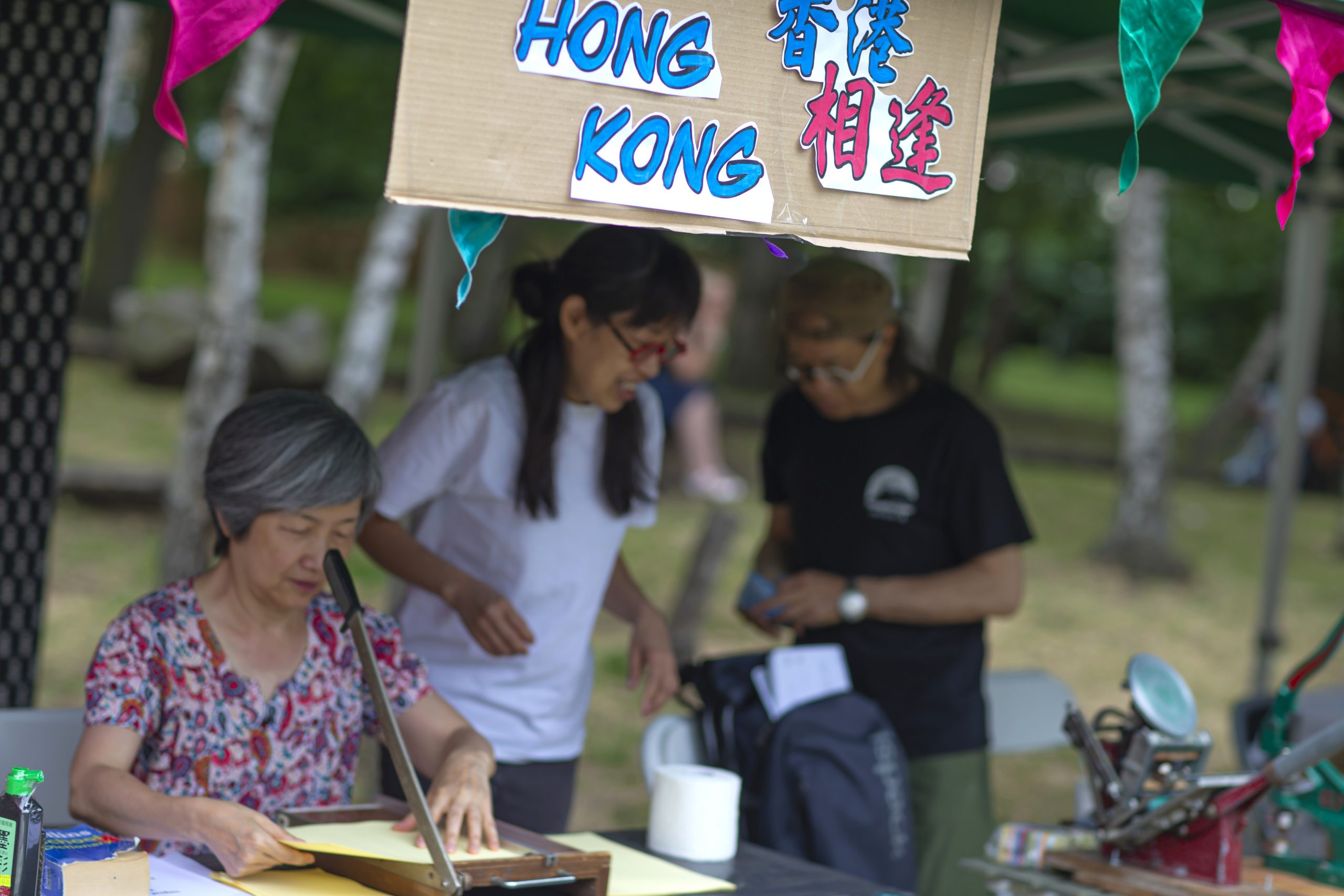 Remembering a glorious Summer in the Park. Three people are at a stall. Above their heads is a sign which reads Hong Kong.