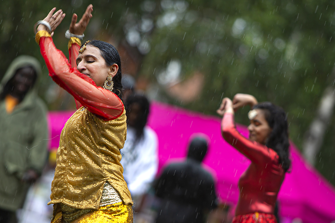 Remembering a glorious Summer in the Park. Two dancers in red and gold have their hands raised. They are outside and it is raining.